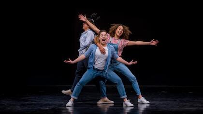 Tre personer dansar på en scen med mörk bakgrund, poserar med utsträckta armar och ben.