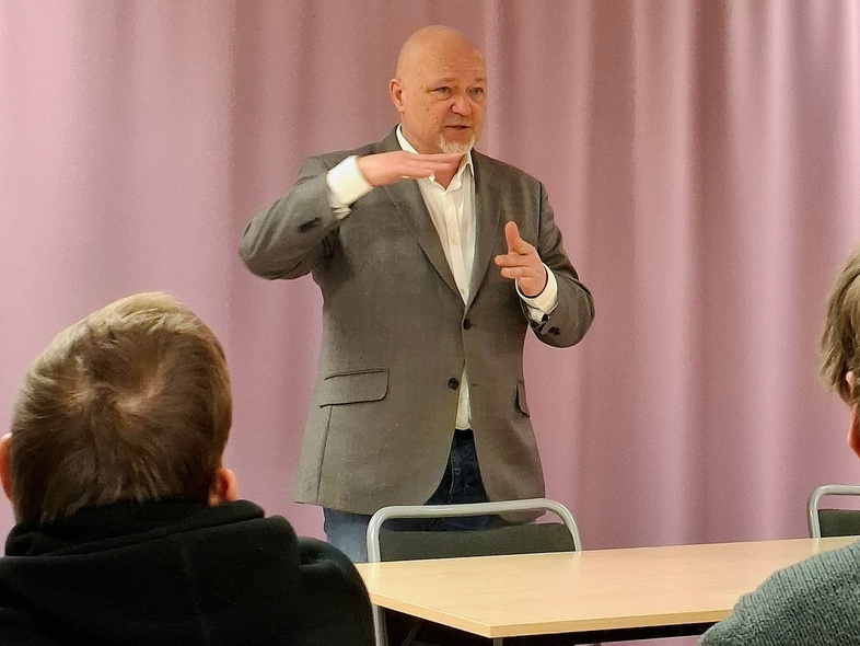 En man i grå kavaj står och talar inför två personer i ett klassrum med lila gardiner.