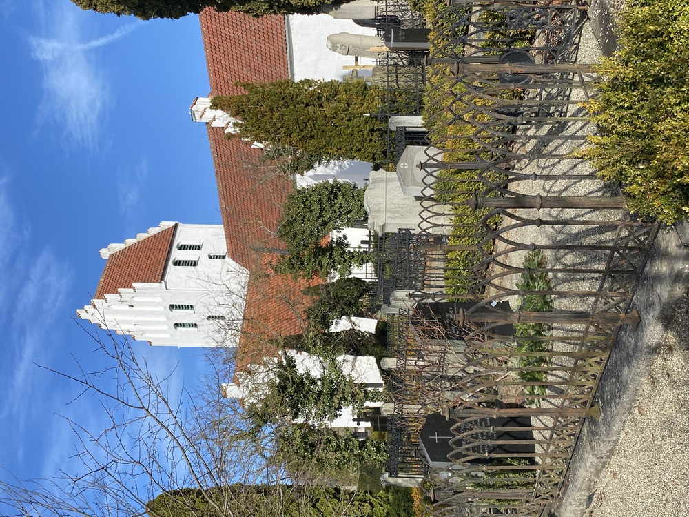 Vit kyrka med rött tak och gravstenar omgiven av järnstaket under blå himmel.