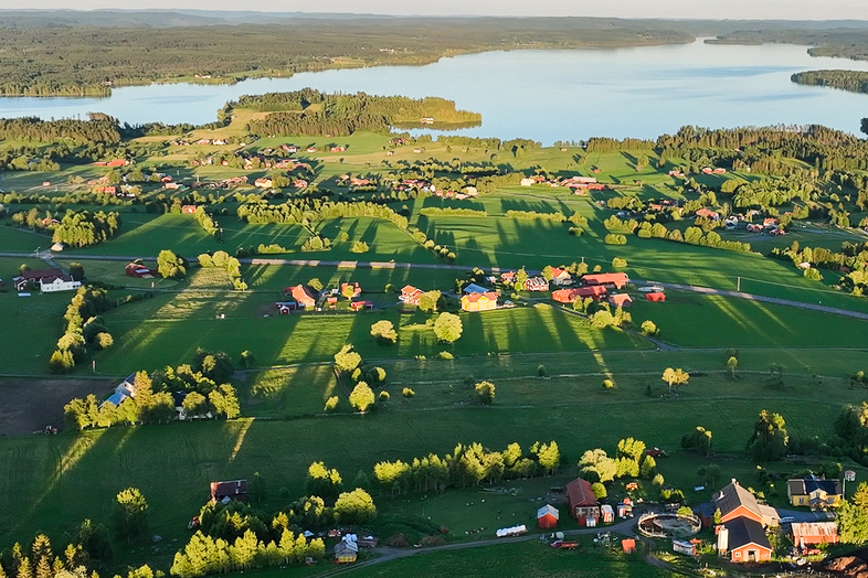 Landskapsvy över gröna fält, gårdar och en sjö i bakgrunden, fotograferat från ovan i kvällssol.