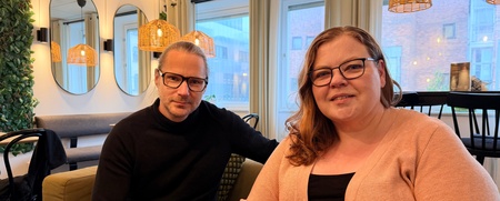 Två personer med glasögon sitter i en modern, mysig kafémiljö med stora fönster och varma lampor.