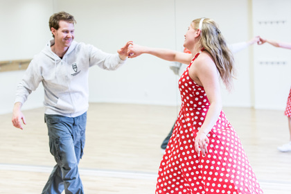 En man och en kvinna dansar tillsammans i en ljus sal. Kvinnan bär en röd prickig klänning.