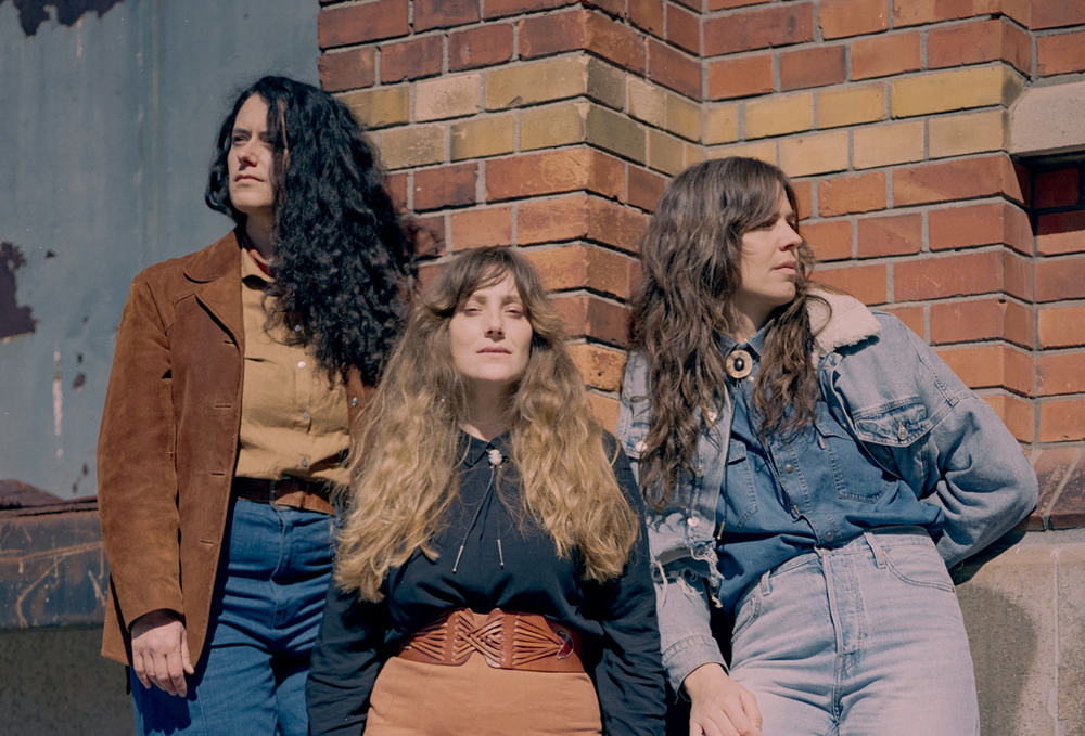 Tre personer står vid en tegelvägg utomhus, klädda i retroinspirerade kläder.