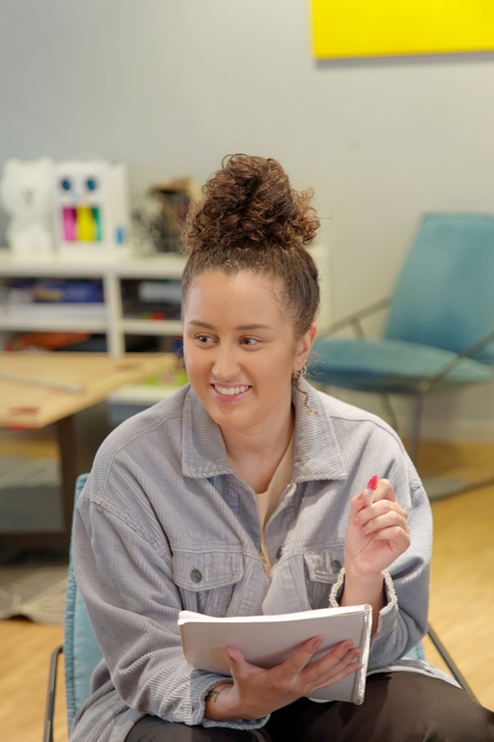 Kvinna med lockigt hår sitter på stol, håller en anteckningsbok och penna, och ler i ett ljust rum.