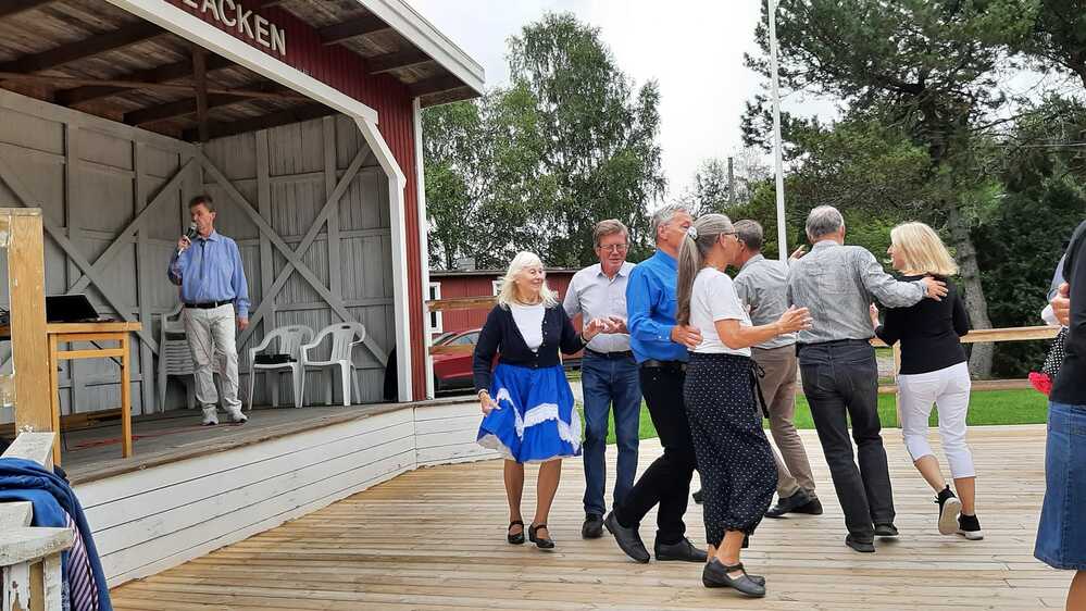 Folk dansar på en träscen utomhus medan en man talar i mikrofon på en upphöjd scen.