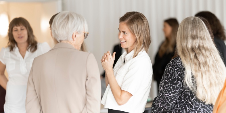 Flera kvinnor samtalar i en ljus inomhusmiljö, några står i fokus och ler mot varandra.