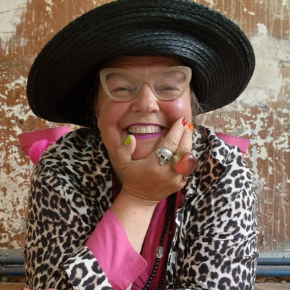 Leende person med stor svart hatt, leopardmönstrad jacka och färgglada accessoarer mot sliten vägg.