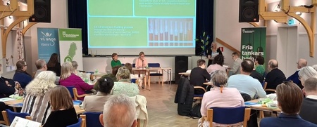 Konferens med deltagare som lyssnar på en presentation i en ljus sal med projektorduk och panel.