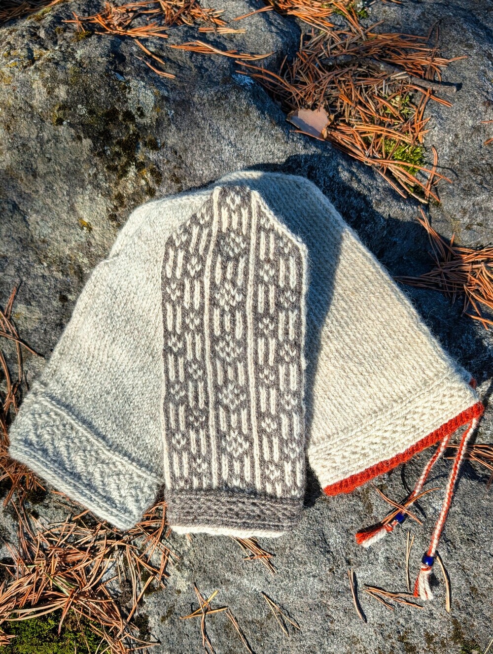 Två stickade vantar i naturfärger ligger på en sten med tallbarr omkring.