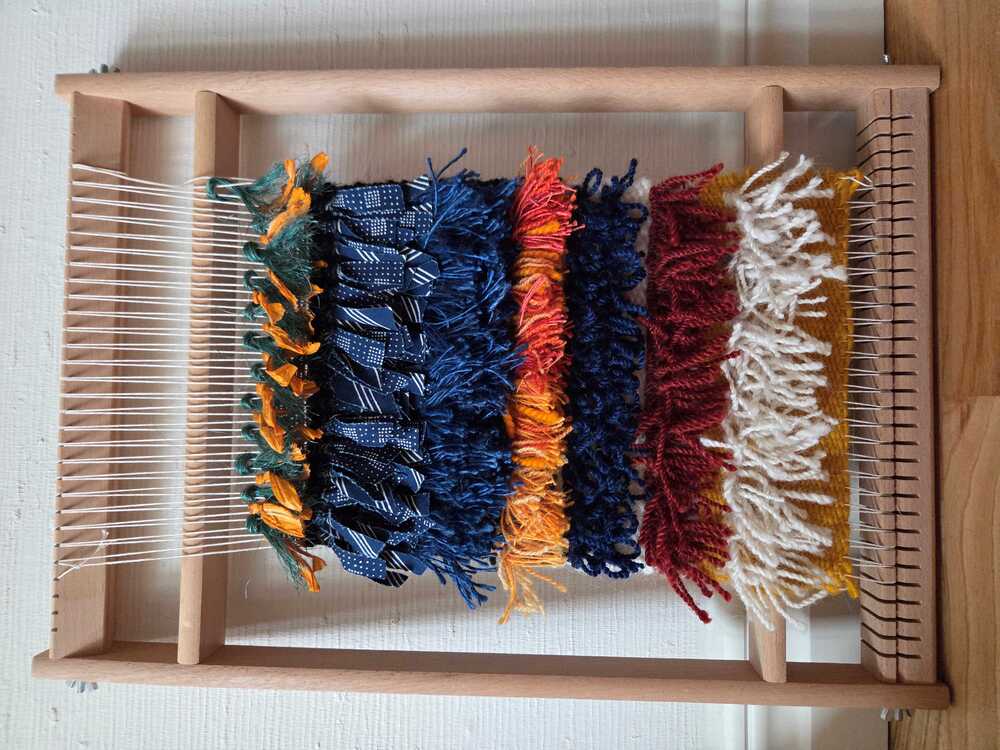 En vävram med färgglada garntrådar i lager av blått, orange, vinrött och vitt mot en vit vägg.