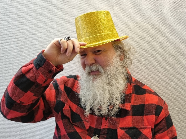 Person med grått skägg och rutig skjorta bär en glittrig gul hatt och håller i hatten med ena handen.