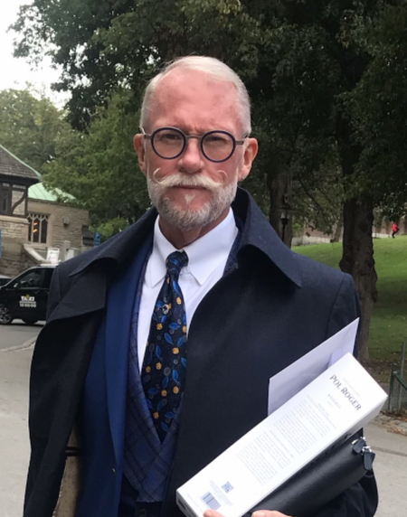 Man i kostym och rock håller en låda och dokument utomhus i parkmiljö.