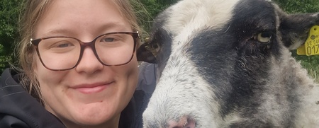 En leende person med glasögon tar en selfie med ett får utomhus, med träd i bakgrunden.