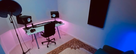 Ett modernt hemmastudio med skrivbord, högtalare, mikrofon, matta och blå soffa, belyst med färgade LED-lampor.