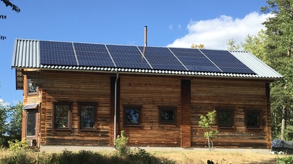 Trähus med solpaneler på taket, omgiven av träd och blå himmel.