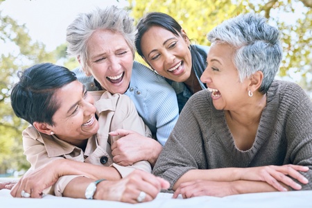 Fyra glada kvinnor i medelåldern skrattar tillsammans utomhus en solig dag.