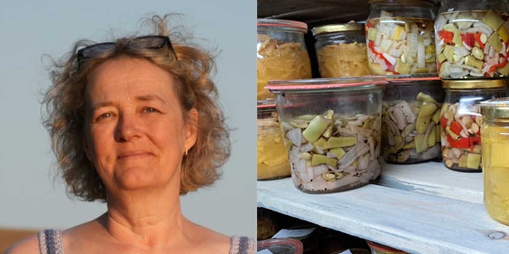 Kvinna med lockigt hår står bredvid hyllor fyllda med inlagda grönsaker i glasburkar.