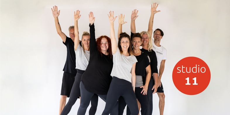 En grupp glada vuxna i träningskläder poserar med armarna uppsträckta framför en vit bakgrund.