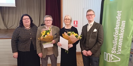 Fyra personer står inomhus, två håller blombuketter och diplom, bredvid en grön rollup med texten 'Studieförbundet Vuxenskolan'.