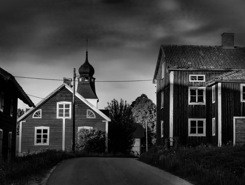 Svartvit bild av en landsväg som leder mellan två gamla trähus, ett med kyrktorn, under dramatisk himmel.