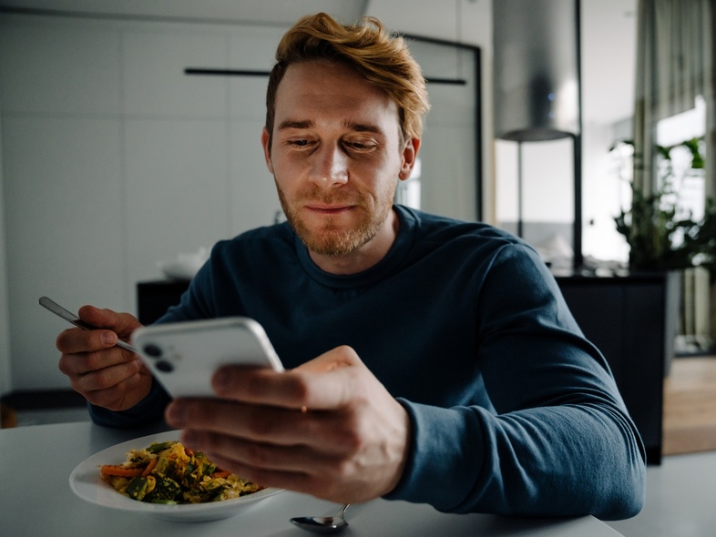 Man sitter vid ett bord, äter mat och tittar på sin mobiltelefon i ett modernt hem.