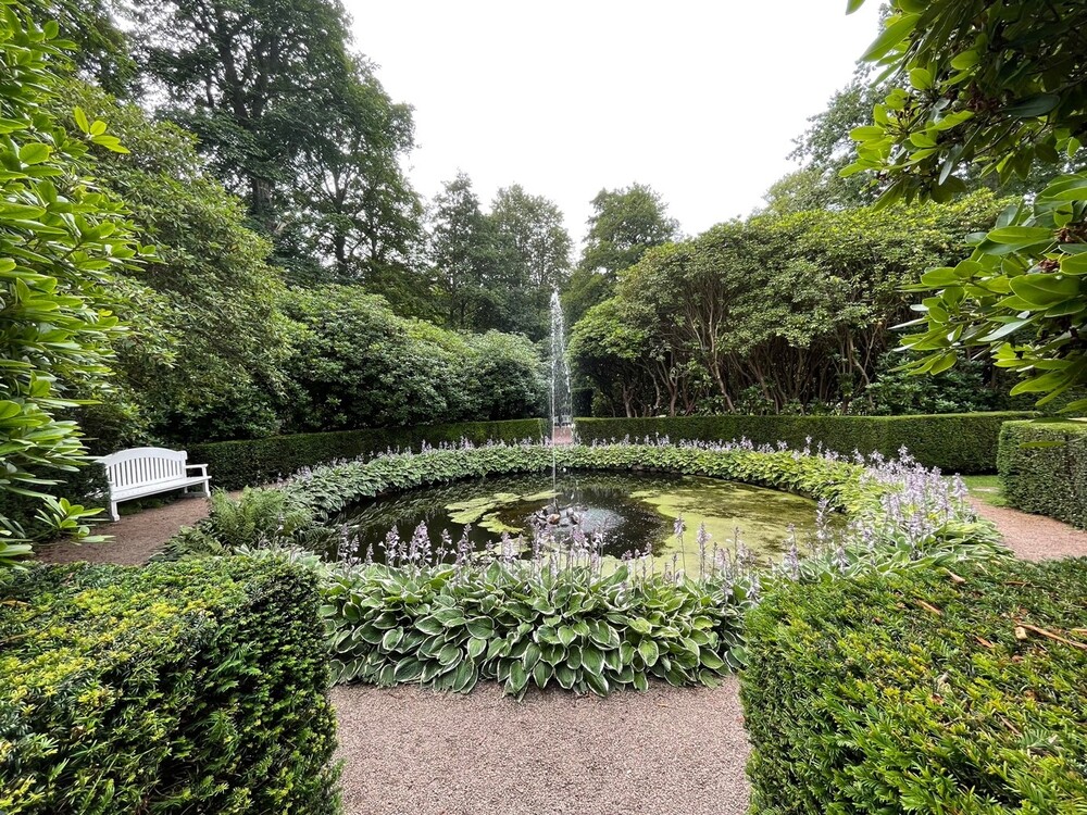 Rund damm med fontän i mitten av en grönskande trädgård, omgiven av buskar och en vit bänk.