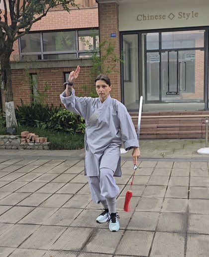 Person i grå traditionell dräkt utför en svärdform utomhus framför en byggnad med skylten 'Chinese Style'.