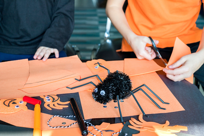 Två personer gör Halloween-pyssel med orange papper, sax och spindeldekoration på ett bord.