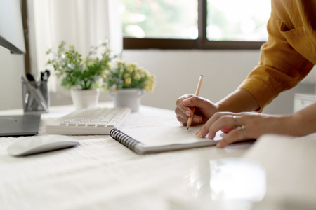 En person skriver i en anteckningsbok vid ett ljust skrivbord med dator, växter och fönster i bakgrunden.