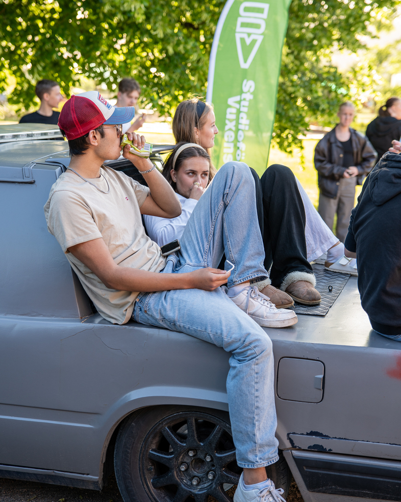 Två unga personer sitter avslappnat på flaket av en bil under ett utomhusevenemang.