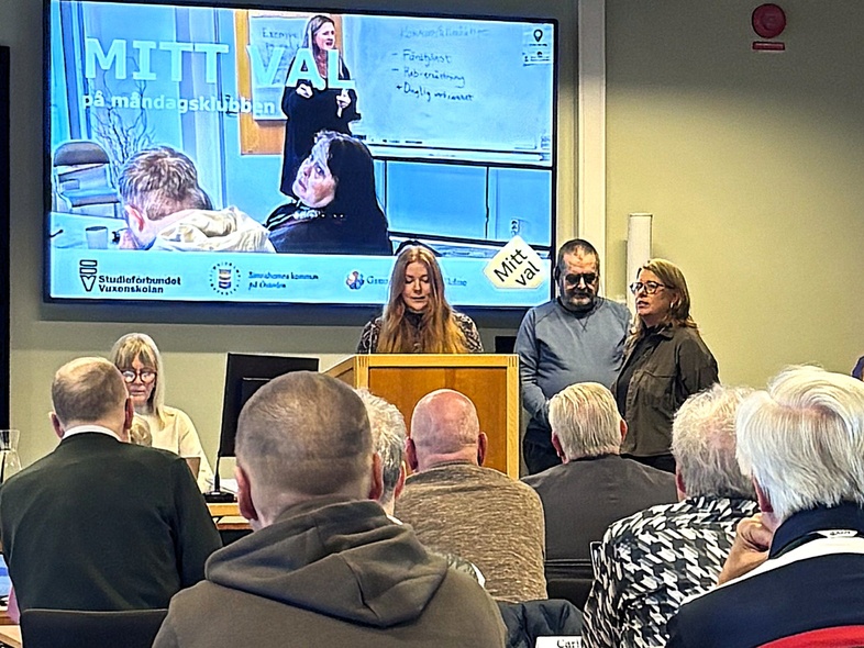 Möte eller presentation i konferensrum med flera personer, en talare vid podiet och publik som lyssnar.