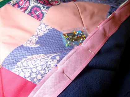Närbild av ett lapptäcke med rosa kantband, fastnålat med knappnålar och en ask med fler nålar ovanpå.