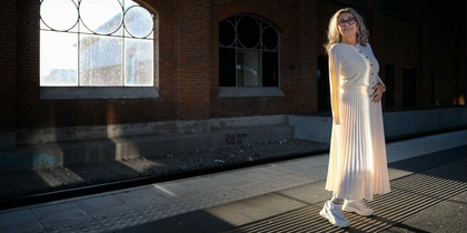 Kvinna i ljus kjol och tröja står på en perrong med tegelvägg och stora fönster i bakgrunden.