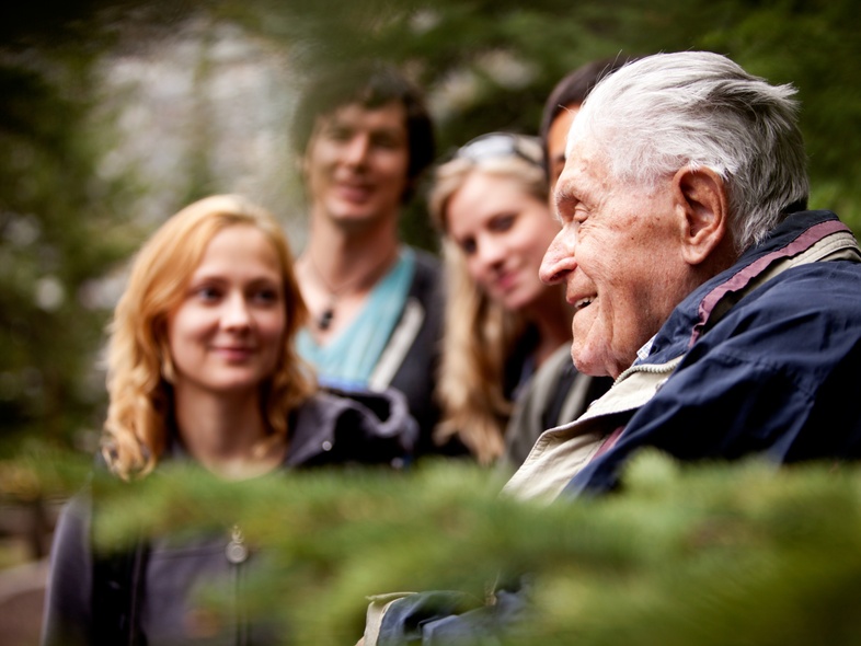 En äldre man pratar med en grupp yngre vuxna utomhus i en grönskande skogsmiljö.