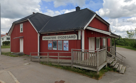 Röd träbyggnad med skylt 'Högsäters Bygdegård' och ramp vid entrén, landsbygdsmiljö.