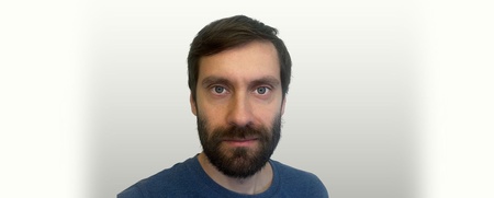 Man med brunt hår och skägg mot ljus bakgrund, tittar rakt mot kameran.
