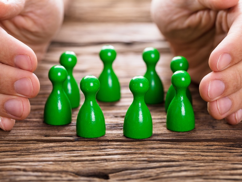 Två händer skyddar en grupp gröna spelpjäser på ett träbord.