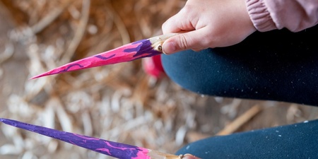 Ett barn håller två färgglada, målade träpinnar med spetsiga ändar, sittande på marken.