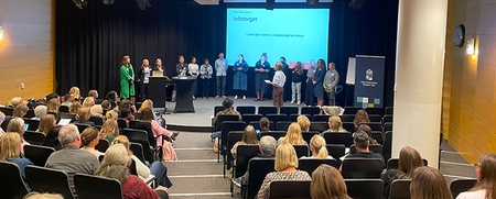 Sittande publik och ett antal personer på scen. Konferensmiljö. 