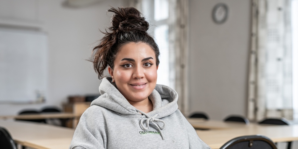 Ung kvinna med mörkt hår i knut och grå hoodie sitter leende i ett klassrum med tomma stolar och bord.