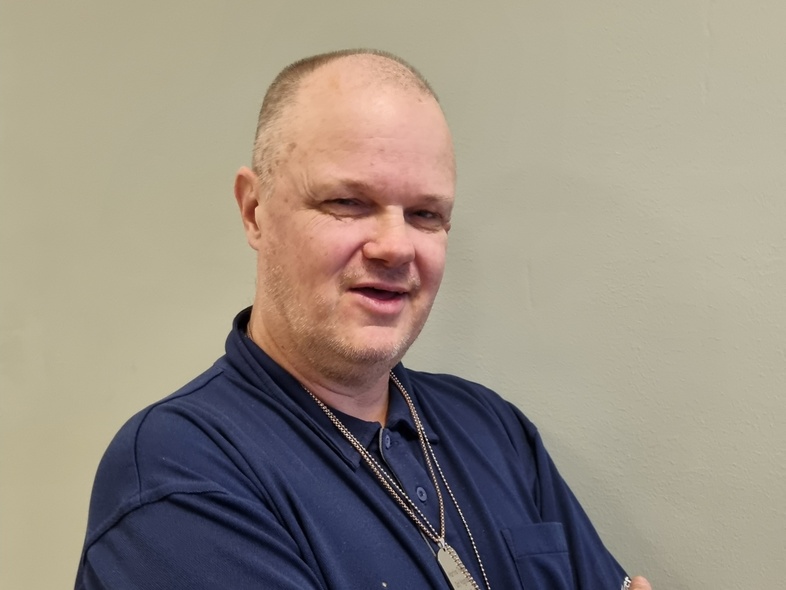 Person i mörkblå pikétröja står med armarna i kors mot en ljus vägg. Ansiktet är suddat.