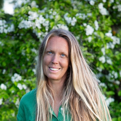 Kvinna med långt blont hår står utomhus framför ett blommande träd, bär grön skjorta och ljusa jeans.