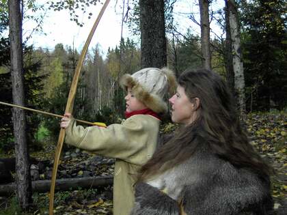 Ett barn och en vuxen i pälsliknande kläder står i en skog. Barnet siktar med en långbåge, den vuxna tittar på.