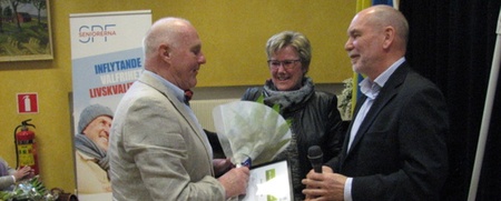 Tre personer står i ett rum, en man överlämnar ett diplom och blommor till en annan man, en kvinna står bredvid och ler.