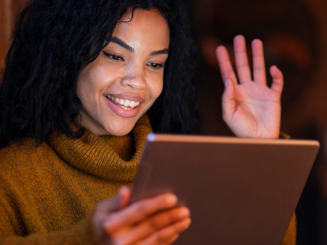 Kvinna med lockigt hår vinkar och ler mot en surfplatta i ett varmt upplyst rum.