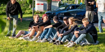 En grupp ungdomar sitter på gräset i solen, några vuxna står i bakgrunden vid bilar.