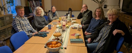 En grupp vuxna sitter runt ett konferensbord med fika, böcker och frukt i ett mötesrum.