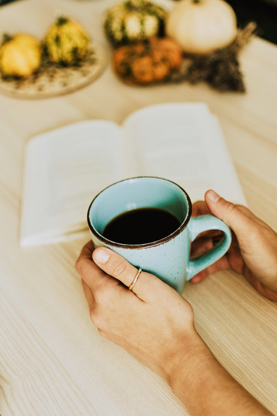 En person håller en blå mugg med kaffe framför en öppen bok på ett bord med pumpor i bakgrunden.