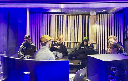 Sex personer sitter i en musikstudio med blå belysning och diskuterar runt ett bord.