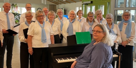 En grupp äldre personer står samlade runt ett piano i en ljus lokal, klädda i vitt och blått.